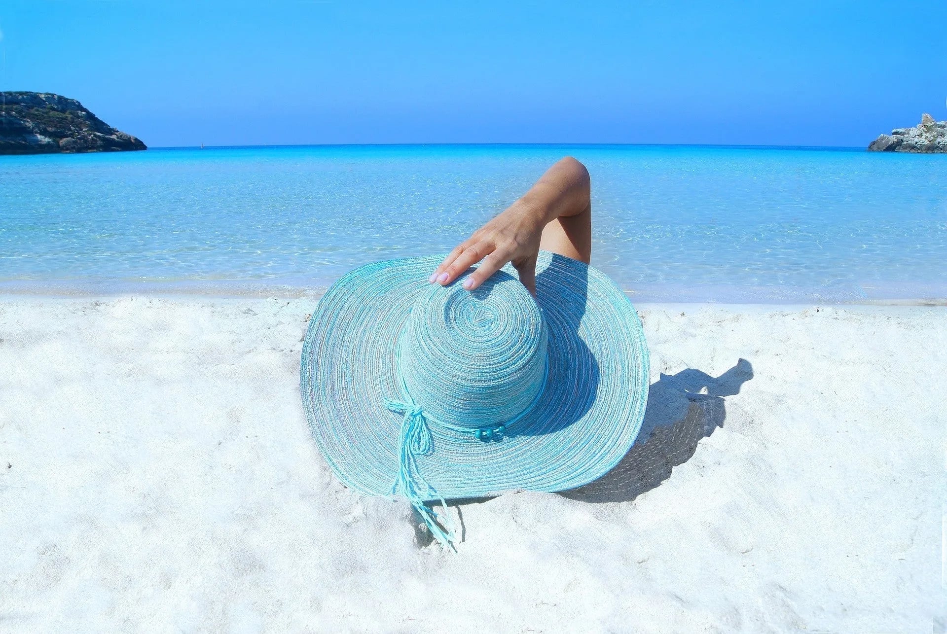 Person lounging on white sand beach under turquoise sun hat